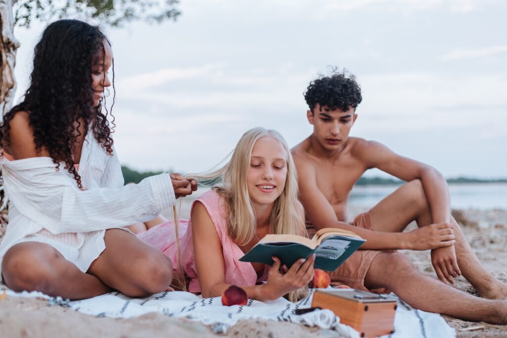 Whoto of two girls and and a guy sitting and reading on a towel on a beach
